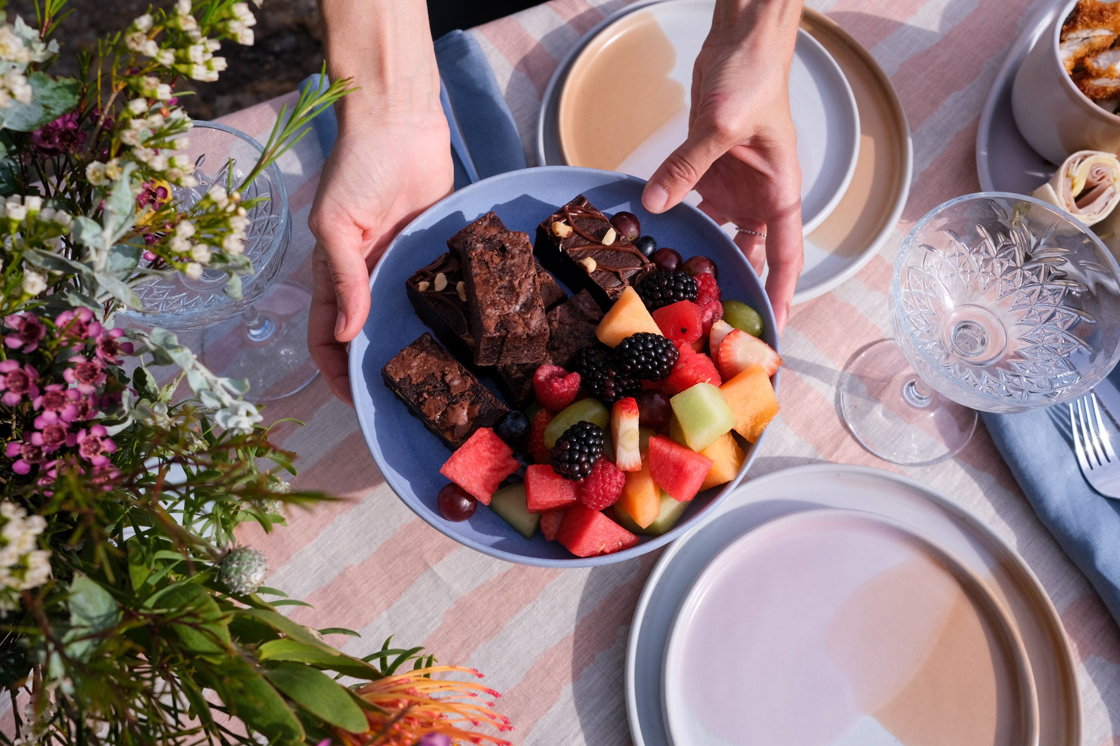 Bright summer outdoor dining table with a handmade blue pottery bowl with fruit and chocolate brownies by Soul Origin and Palinopsia Ceramics dinnerware sets in pink and chocolate