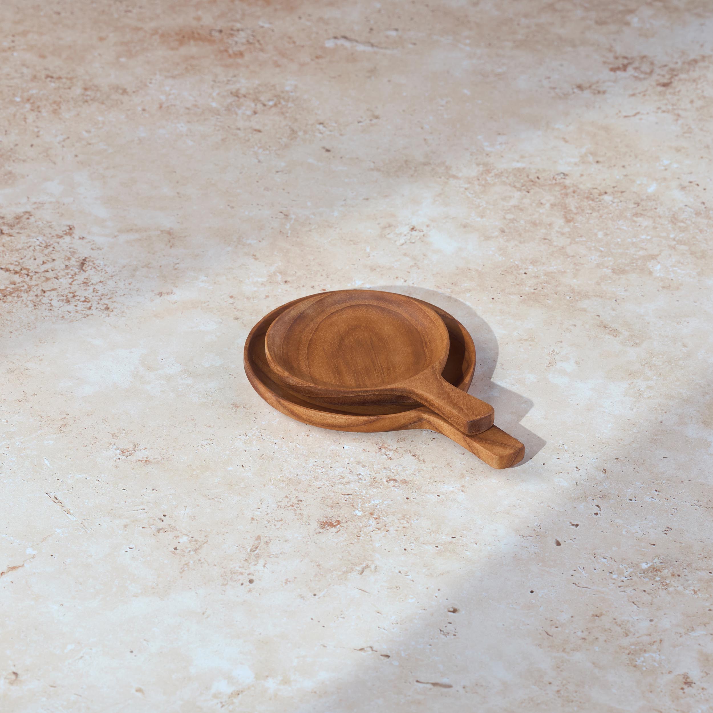 Two circular teak serving dishes with handles of different sizes stacked on a light stone background.