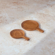 Two circular teak serving dishes with handles, side by side, on a light stone background.