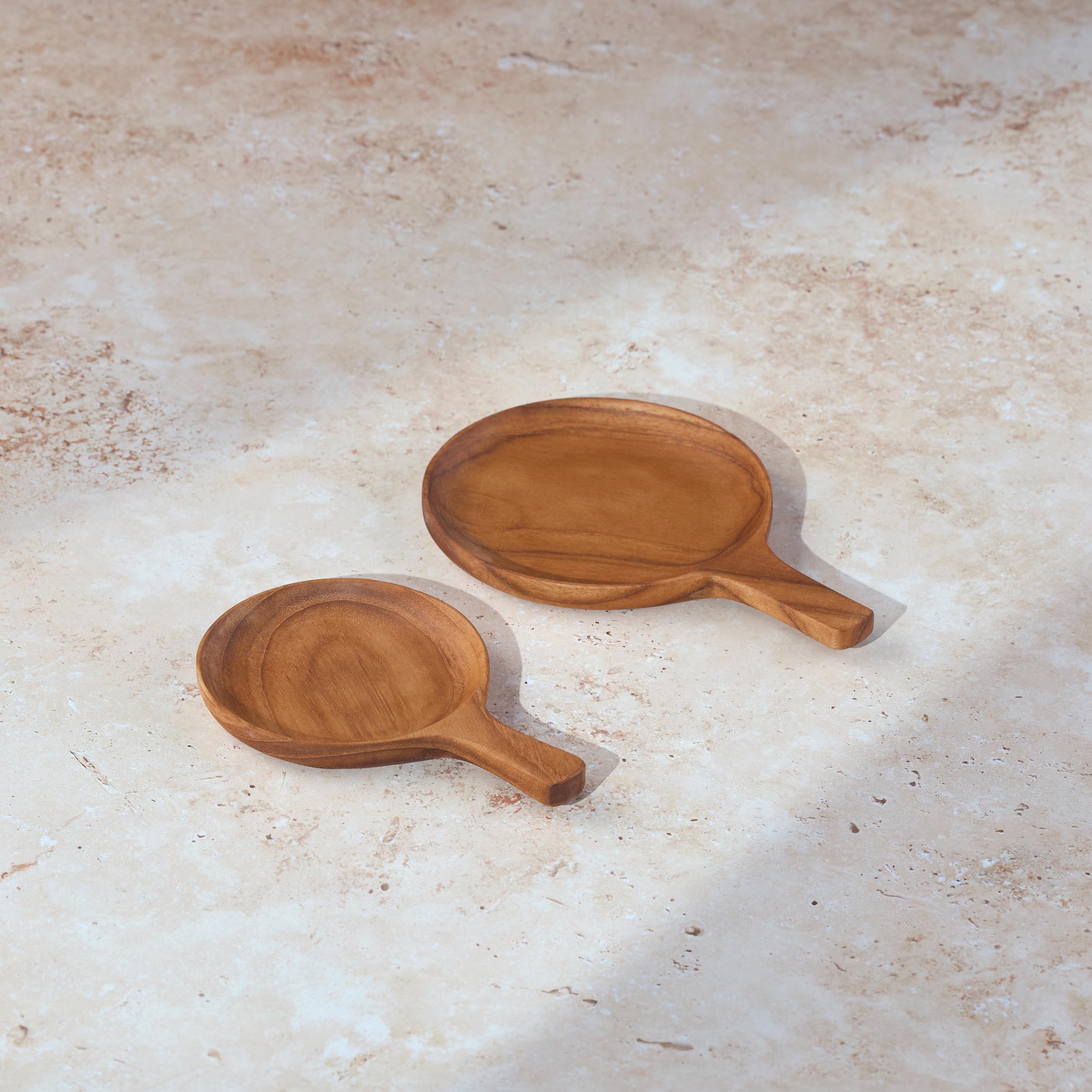 Two circular teak serving dishes with handles, side by side, on a light stone background.