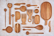 A variety of Palinopsia wooden teak serving dishes, salad servers, bowls, trays, knives, cheese boards in many shapes and sizes on a light stone background.