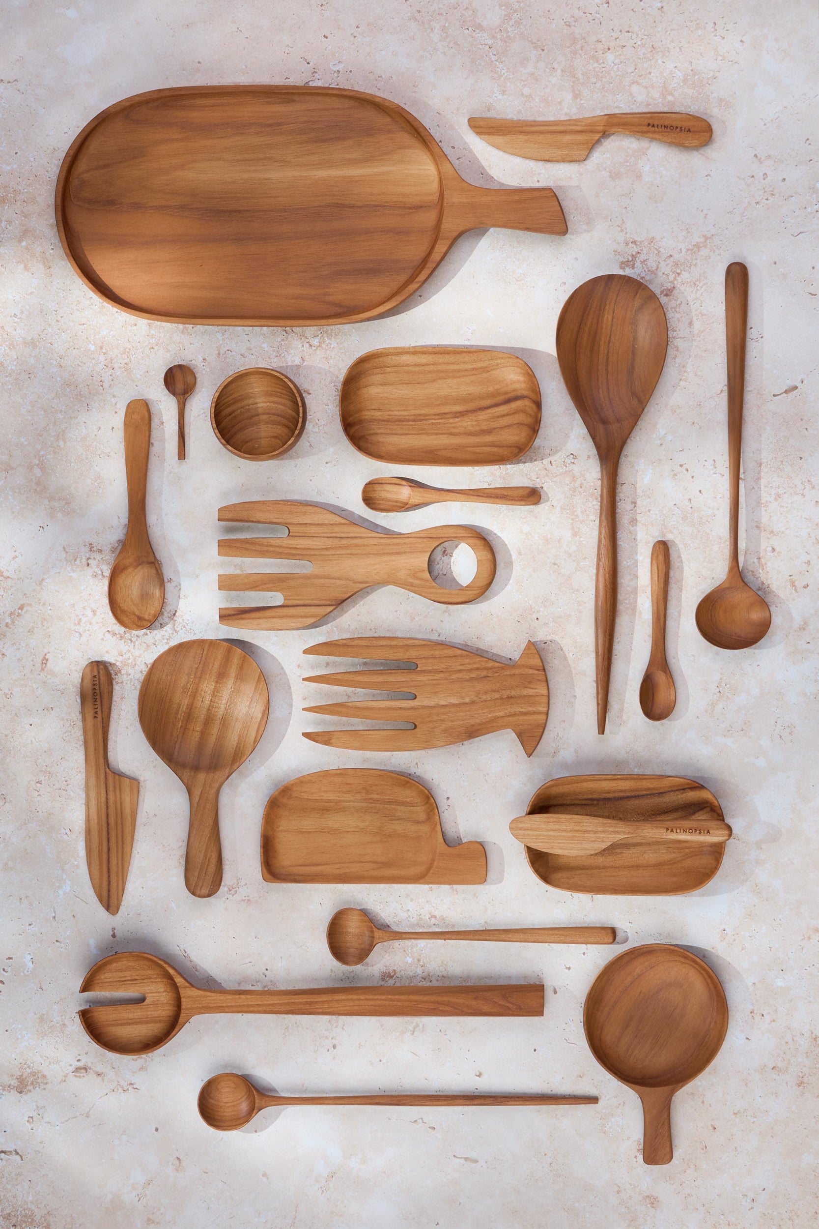 A variety of Palinopsia wooden teak serving dishes, salad servers, bowls, trays, knives, cheese boards in many shapes and sizes on a light stone background.