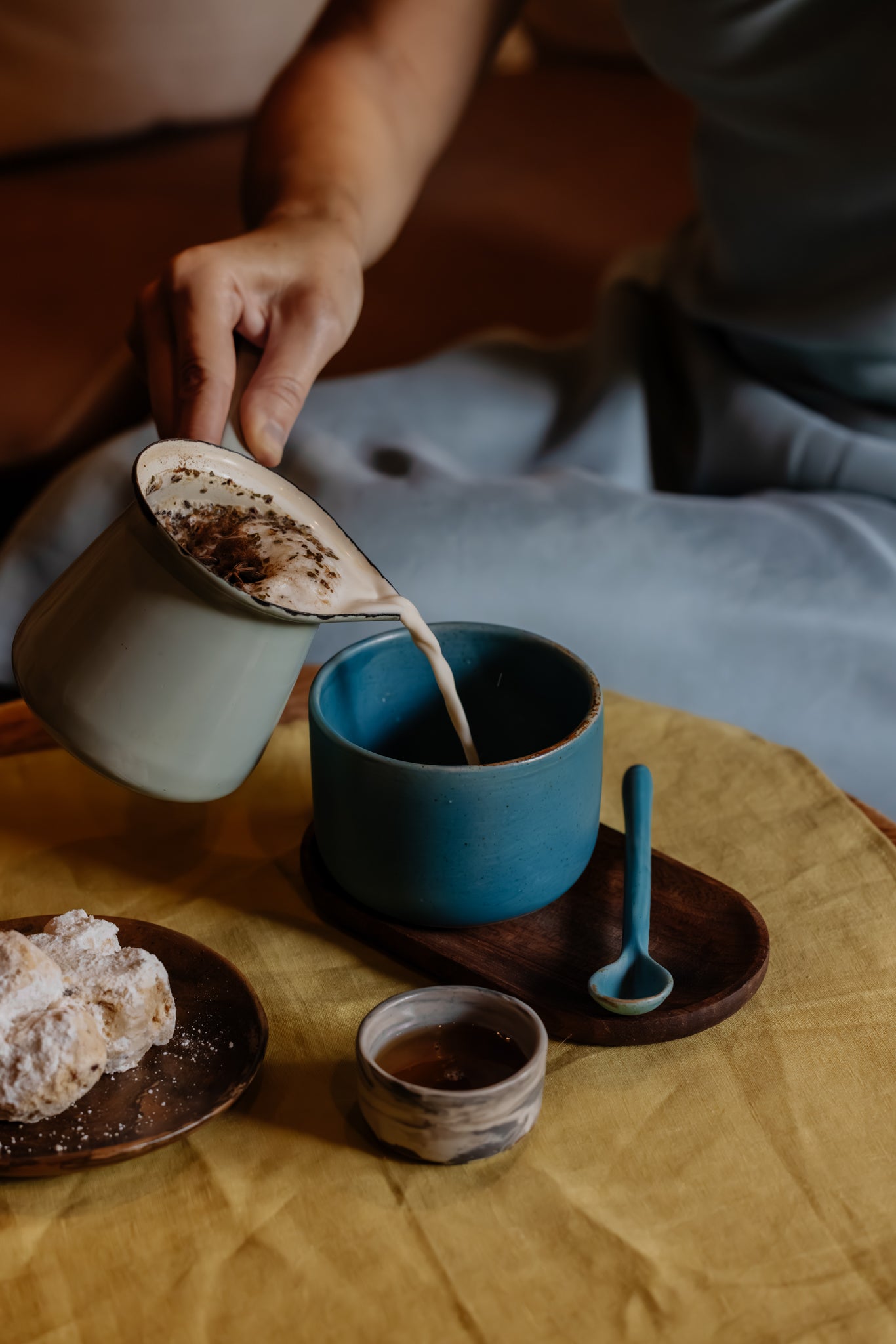 Woman pouring a chai tea with cookies and handmade ceramic teaspoon and honey dish accessories on yellow linen tablecloth
