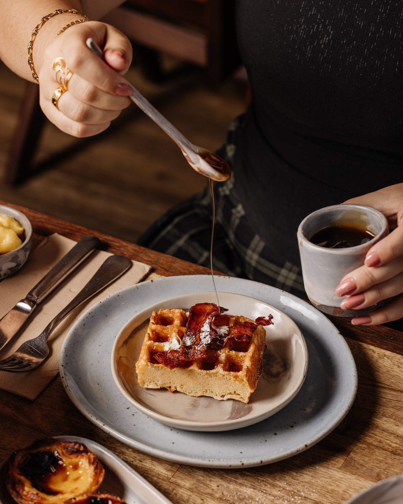 Person pouring syrup onto a bacon waffle with a cup of coffee on a ceramic teaspoon on a table.