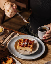Person pouring syrup onto a bacon waffle with a cup of coffee on a ceramic teaspoon on a table.
