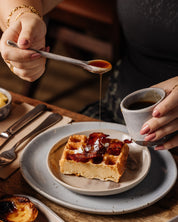Person pouring syrup onto a waffle with bacon and with a ceramic holding a handmade espresso cup 
