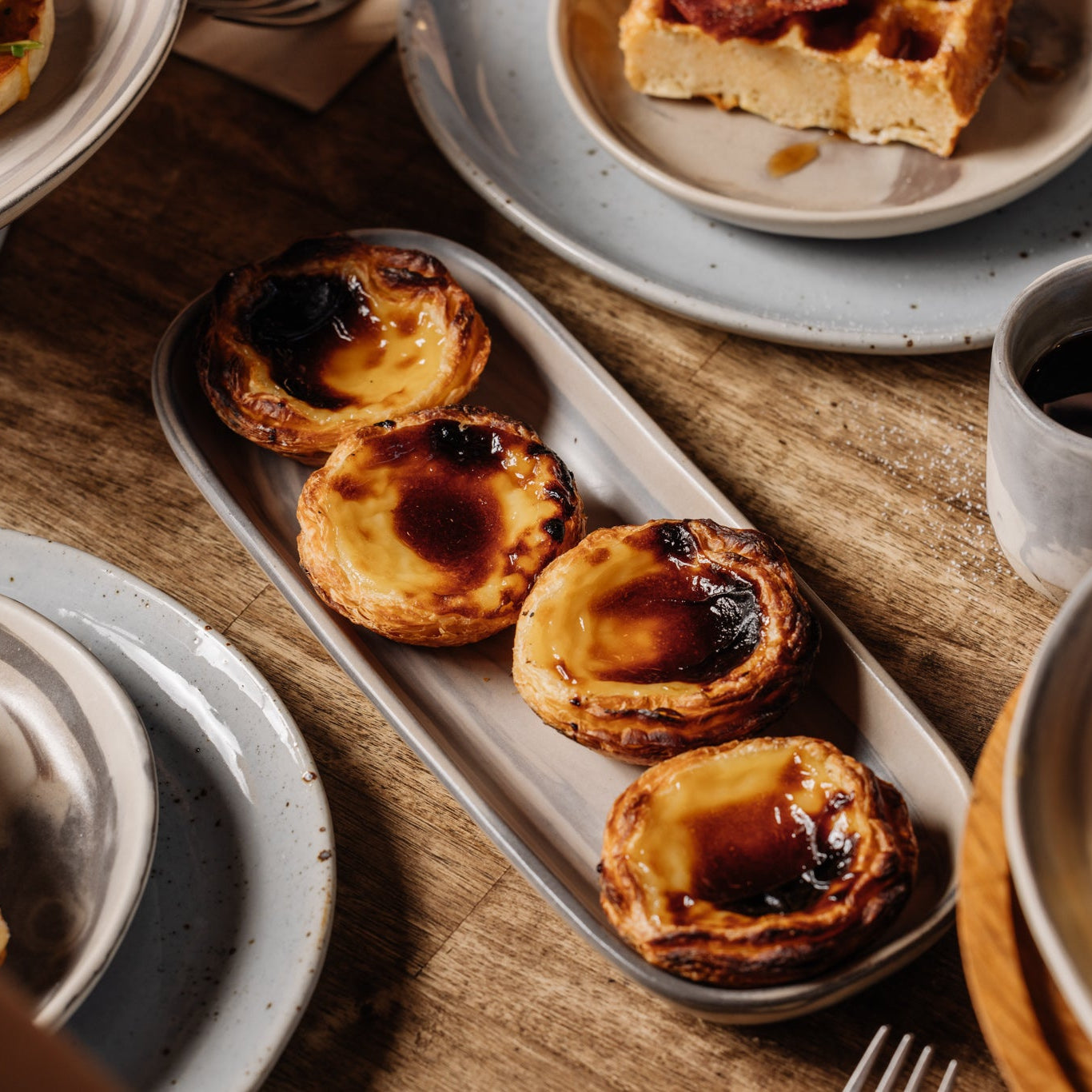 Rectangle ceramic dish with four Portuguese tarts on a breakfast table with a timber table top 