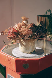 A dried floral arrangement in a small beige pot, displayed on a round marble plinth atop an orange-red vintage stand-up ceramic wheel. The bouquet features dusty pink, brown, and muted red botanicals with delicate hanging strands in Palinopsia Ceramic store in Newcastle. 