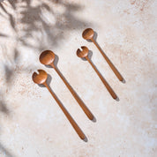 Bird’s-eye view of two sets, one large and one small, of large teak wooden salad servers on a light stone background.