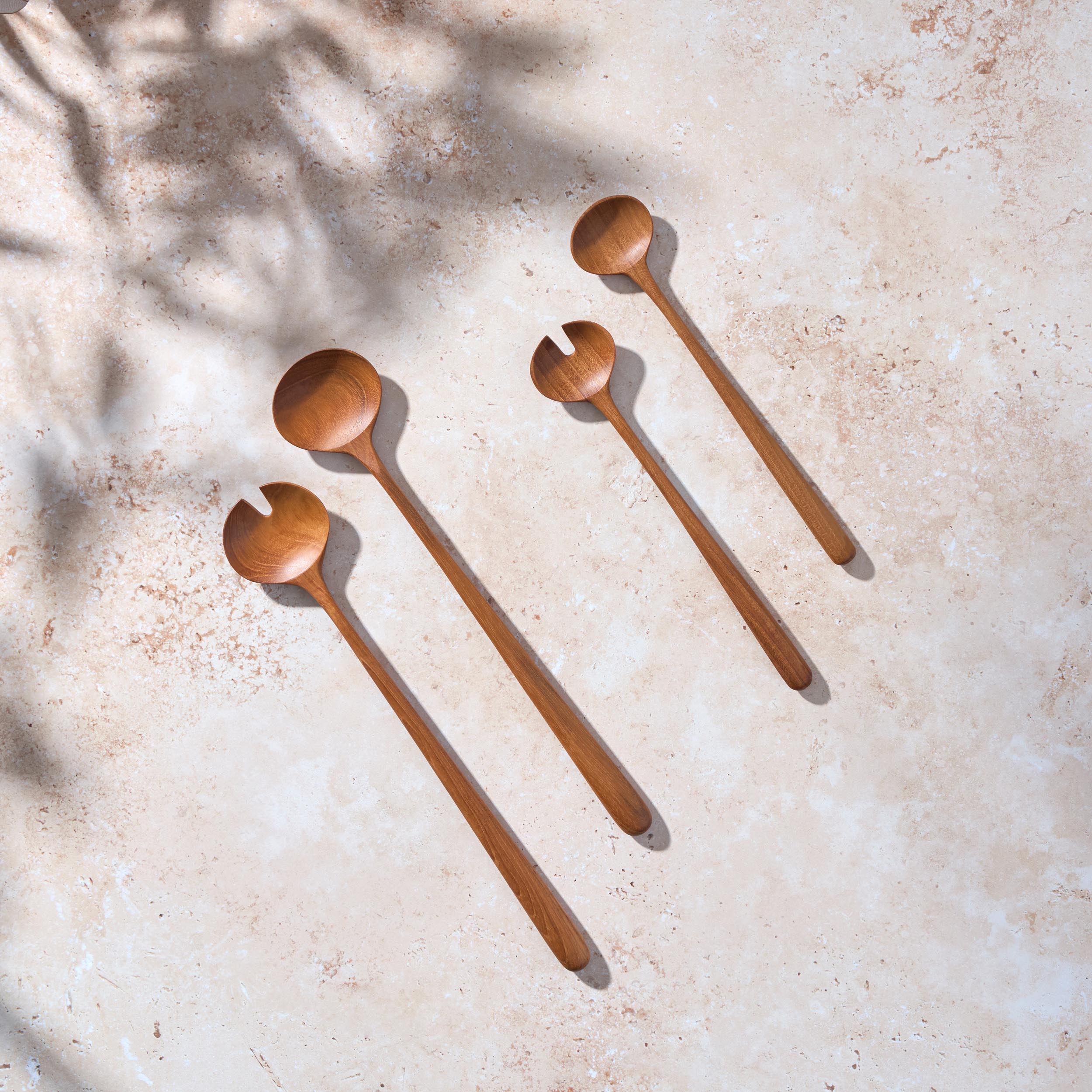 Bird’s-eye view of two sets, one large and one small, of large teak wooden salad servers on a light stone background.