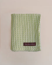 A light green textured cotton towel vertical waffle-like stripes and a small brown PALINOPSIA brand label near the bottom, displayed flat against a warm beige background.