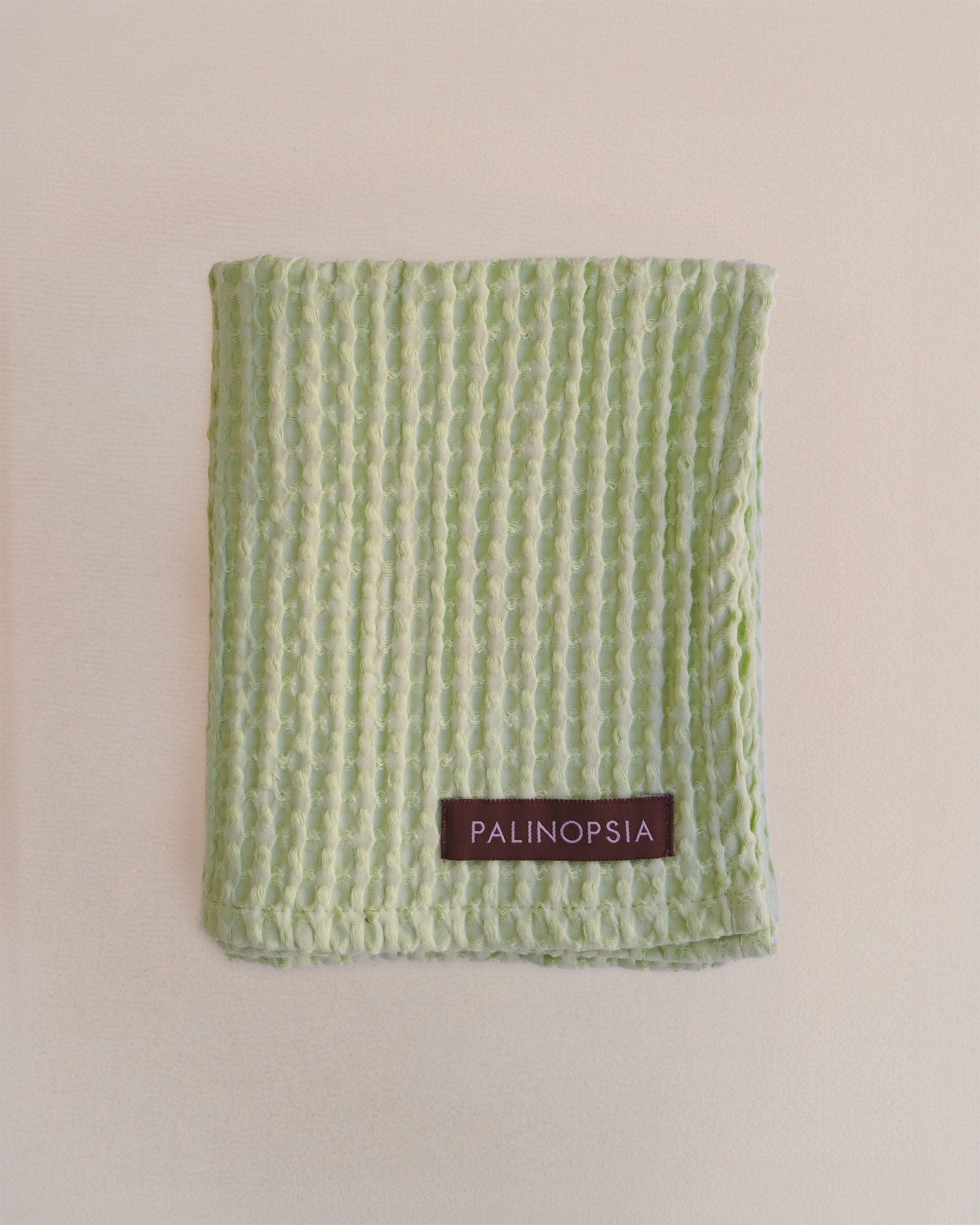 A light green textured cotton towel vertical waffle-like stripes and a small brown PALINOPSIA brand label near the bottom, displayed flat against a warm beige background.
