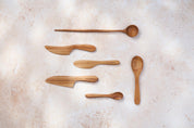 A variety of Palinopsia teak wooden spoons and knives of different shapes and sizes on a light stone background.