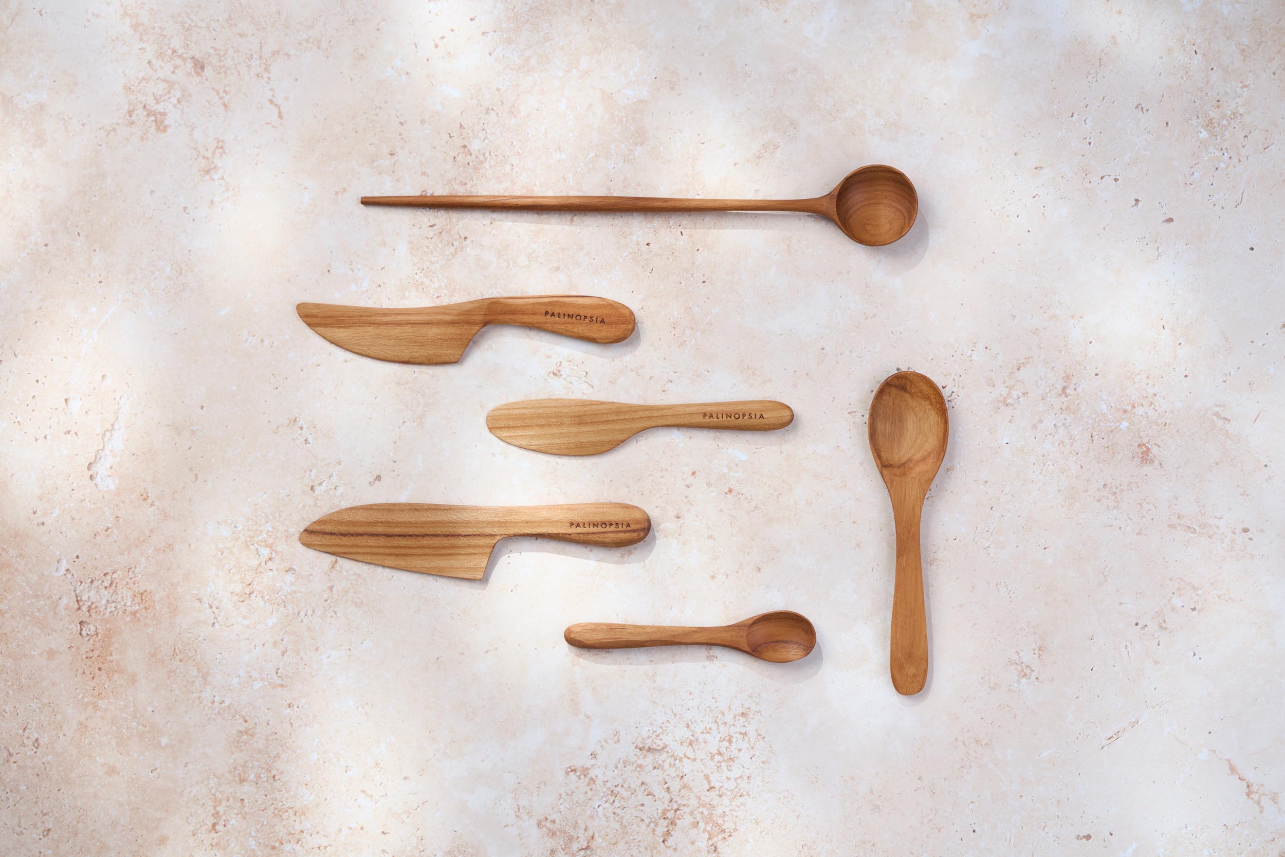 A variety of Palinopsia teak wooden spoons and knives of different shapes and sizes on a light stone background.