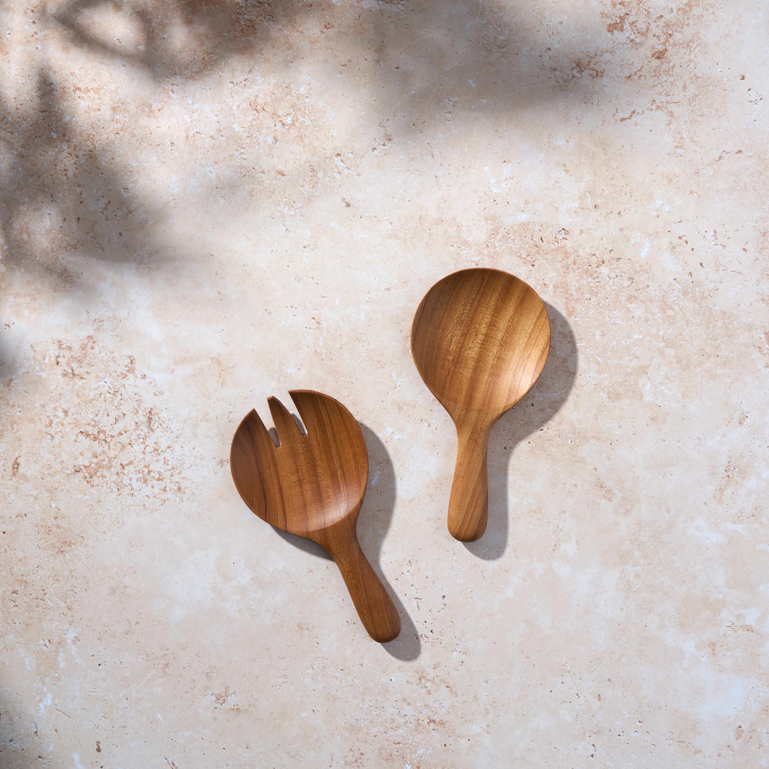 Bird’s-eye view of a short teak wooden salad serving set by Palinopsia on a light stone background.
