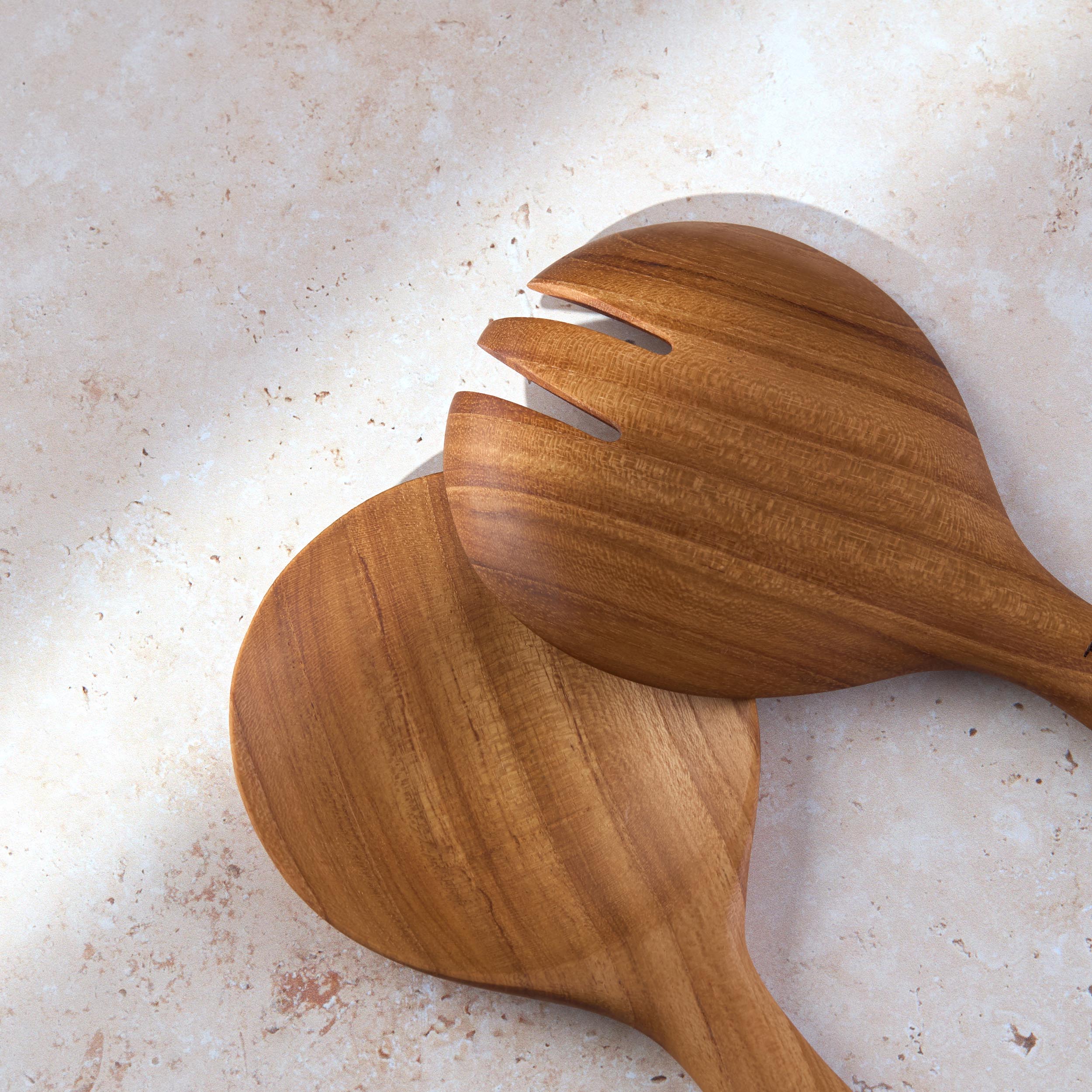 Detailed shot of a short teak wooden salad serving set by Palinopsia on a light stone background.