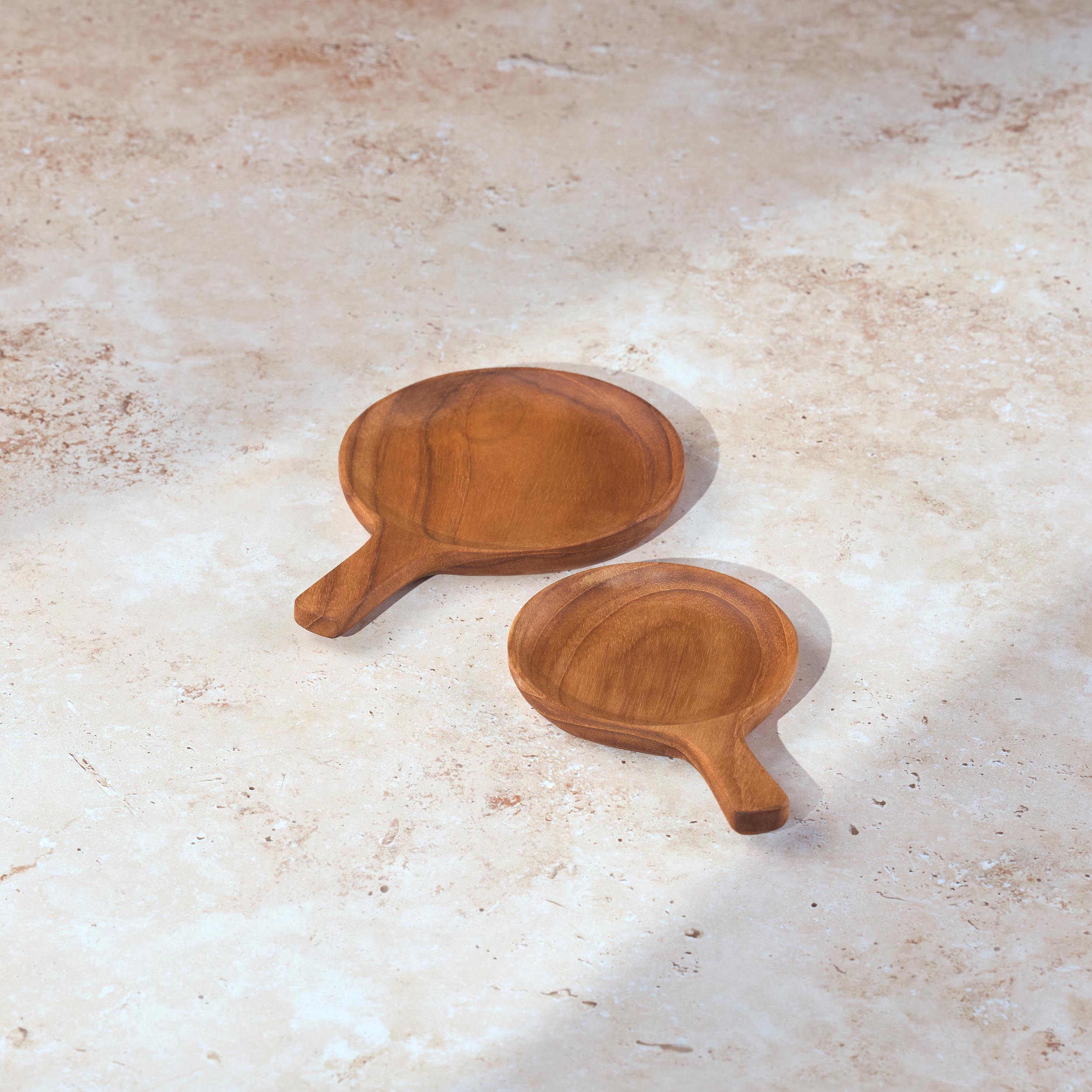Two circular teak serving dishes with handles on a light stone background.