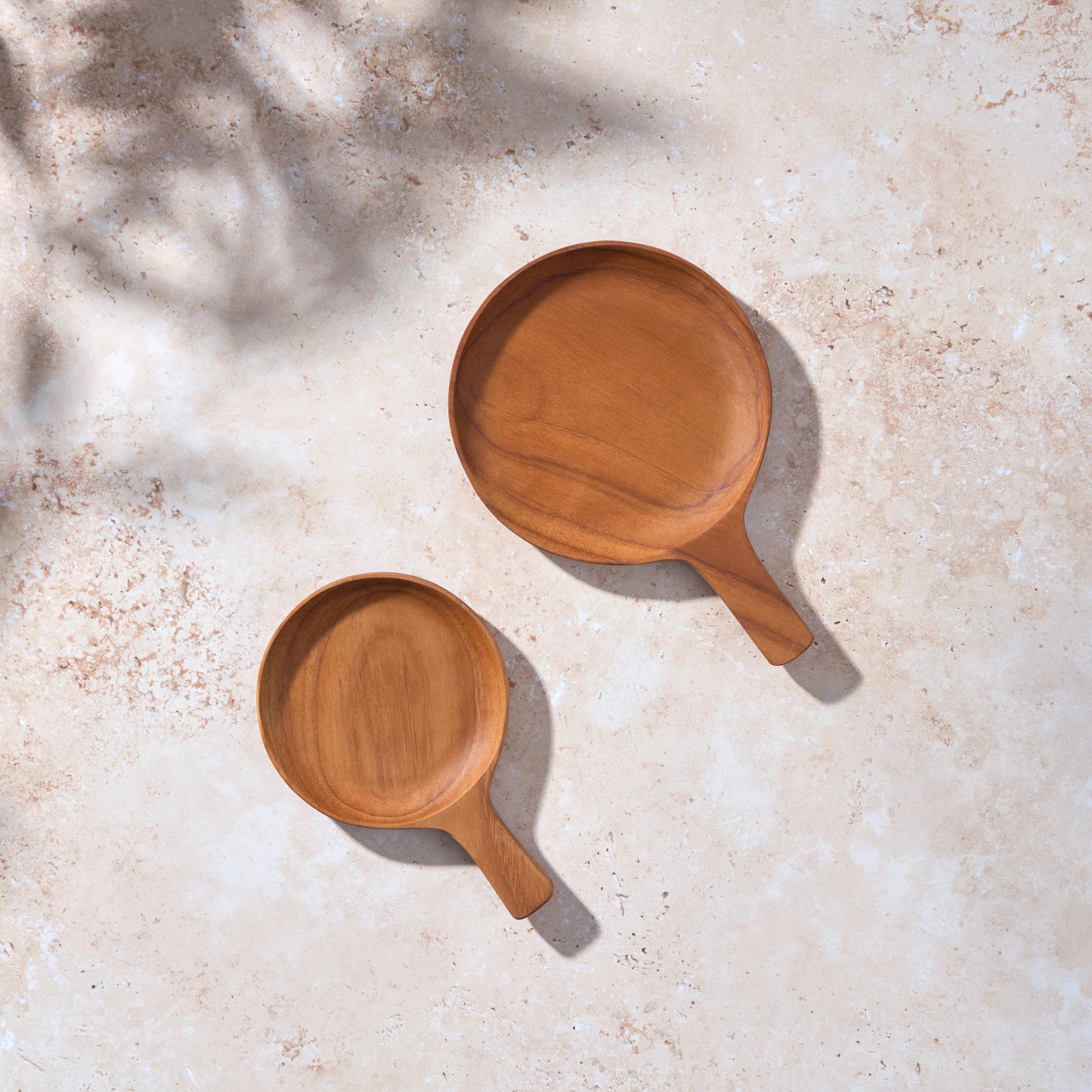 A bird’s-eye view of two circular teak serving dishes with handles on a light stone background.