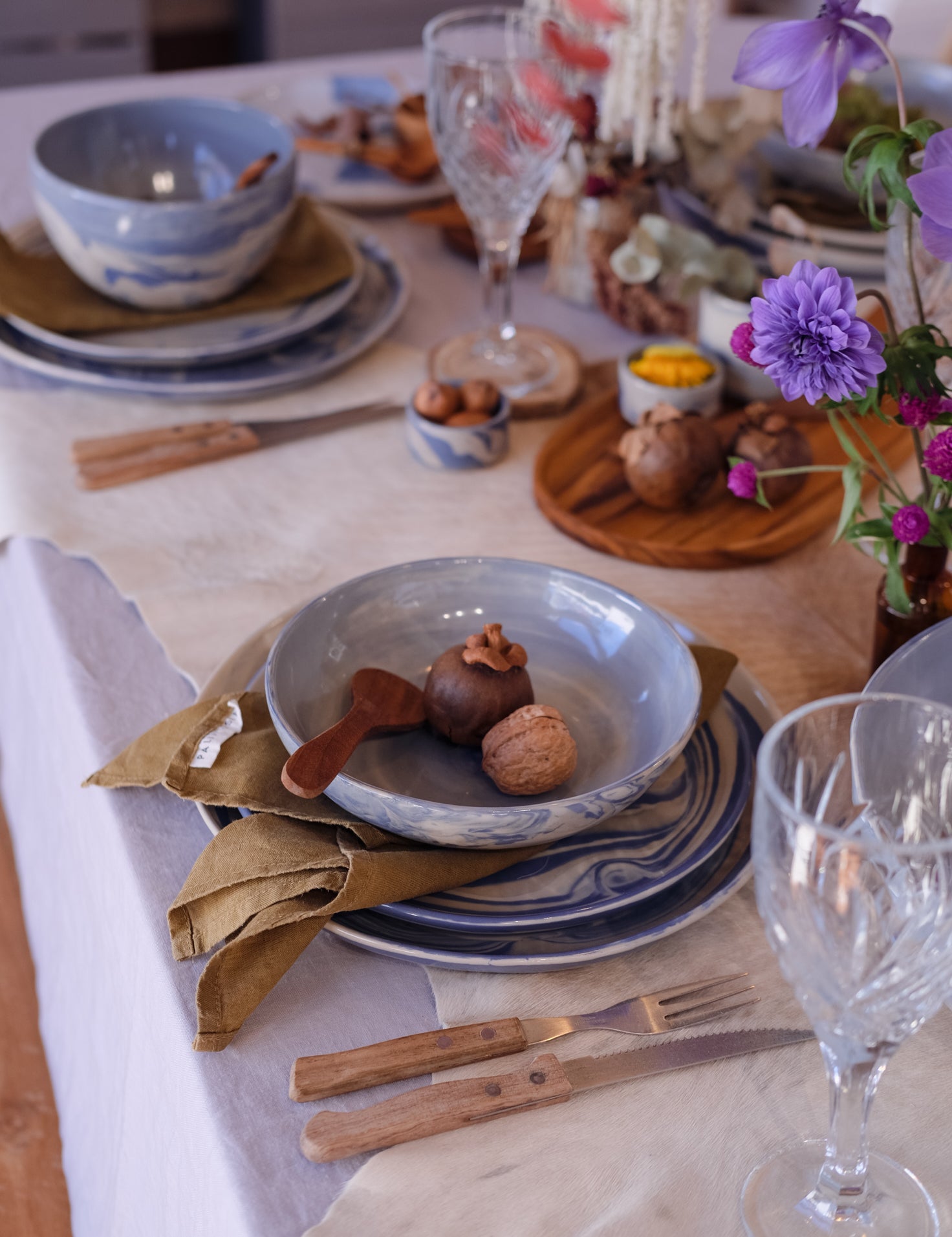 A styled tabletop setting with three piece ceramic dinner set, bowls and plates in blue and white, a marbled blue bowl holding walnuts and a wooden spoon, olive napkins, crystal wine glasses, and purple flowers on a rustic table runner at a Newcastle homewares store.