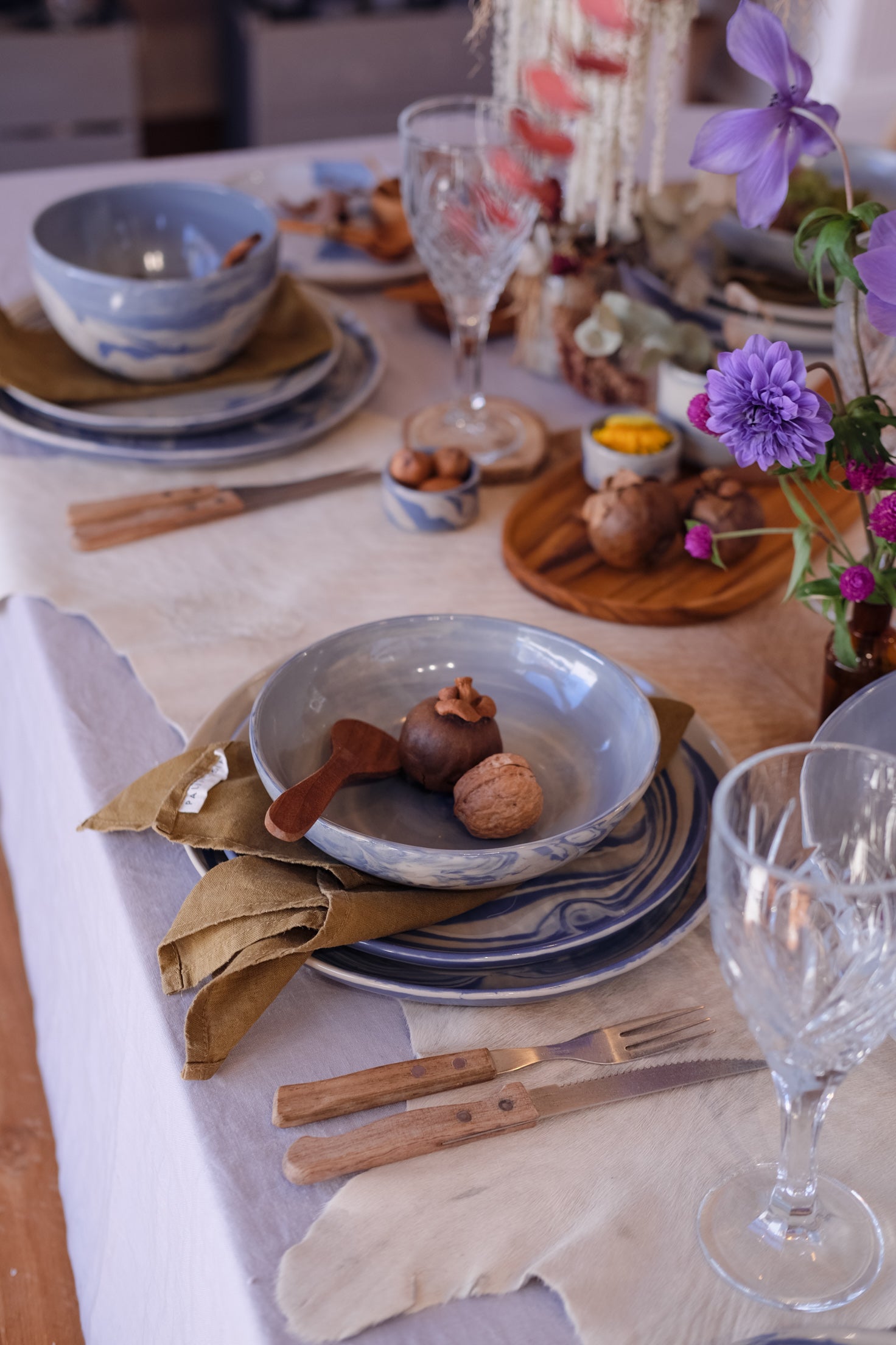 A styled tabletop setting with three piece ceramic dinner set, bowls and plates in blue and white, a marbled blue bowl holding walnuts and a wooden spoon, olive napkins, crystal wine glasses, and purple flowers on a rustic table runner at a Newcastle homewares store.