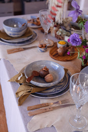 A styled tabletop setting with three piece ceramic dinner set, bowls and plates in blue and white, a marbled blue bowl holding walnuts and a wooden spoon, olive napkins, crystal wine glasses, and purple flowers on a rustic table runner at a Newcastle homewares store.