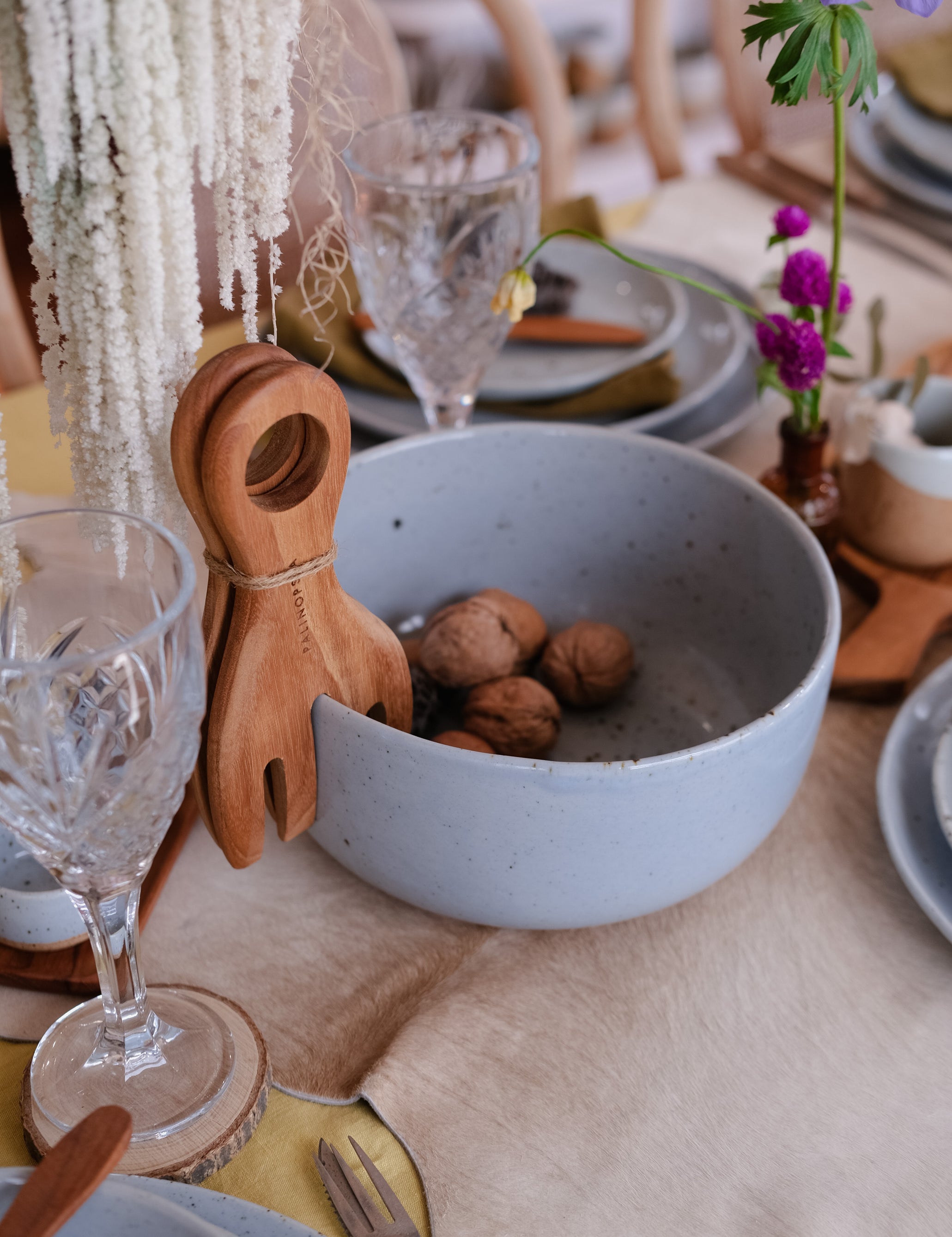 A styled dining table setting featuring a large speckled light blue ceramic fruit bowl filled with walnuts, accompanied by wooden salad servers, clear crystal wine glasses, beige linen placemats, and small floral accents.
