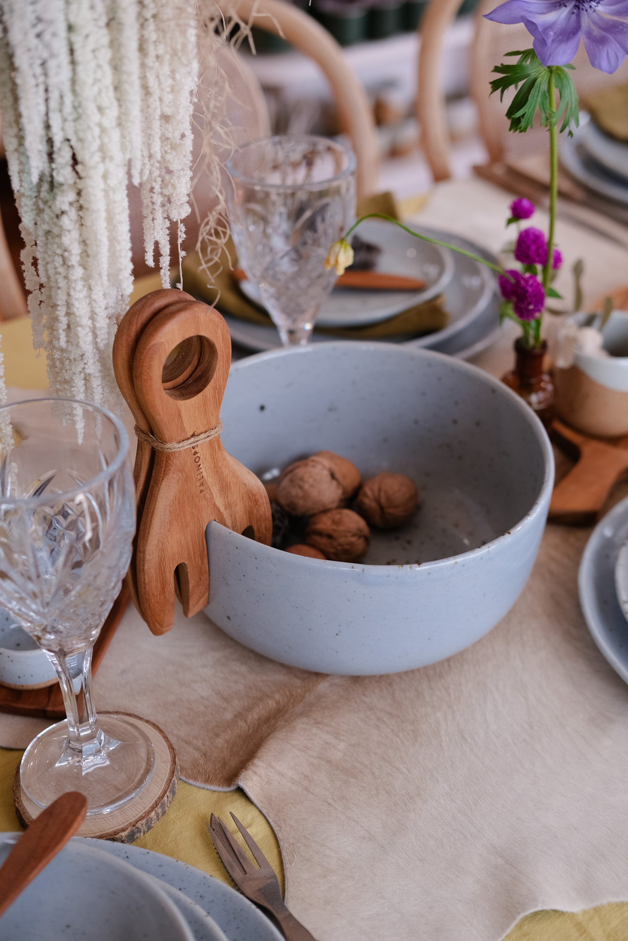 A styled dining table setting featuring a large speckled light blue ceramic fruit bowl filled with walnuts, accompanied by wooden salad servers, clear crystal wine glasses, beige linen placemats, and small floral accents.