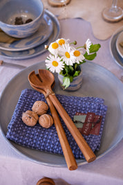 A pair of large teak wooden salad servers on a denim blue waffle tea towel, on a large round ceramic platter. A nicely set dinner table with a light grey tablecloth and blue dinner plates are in the background. 