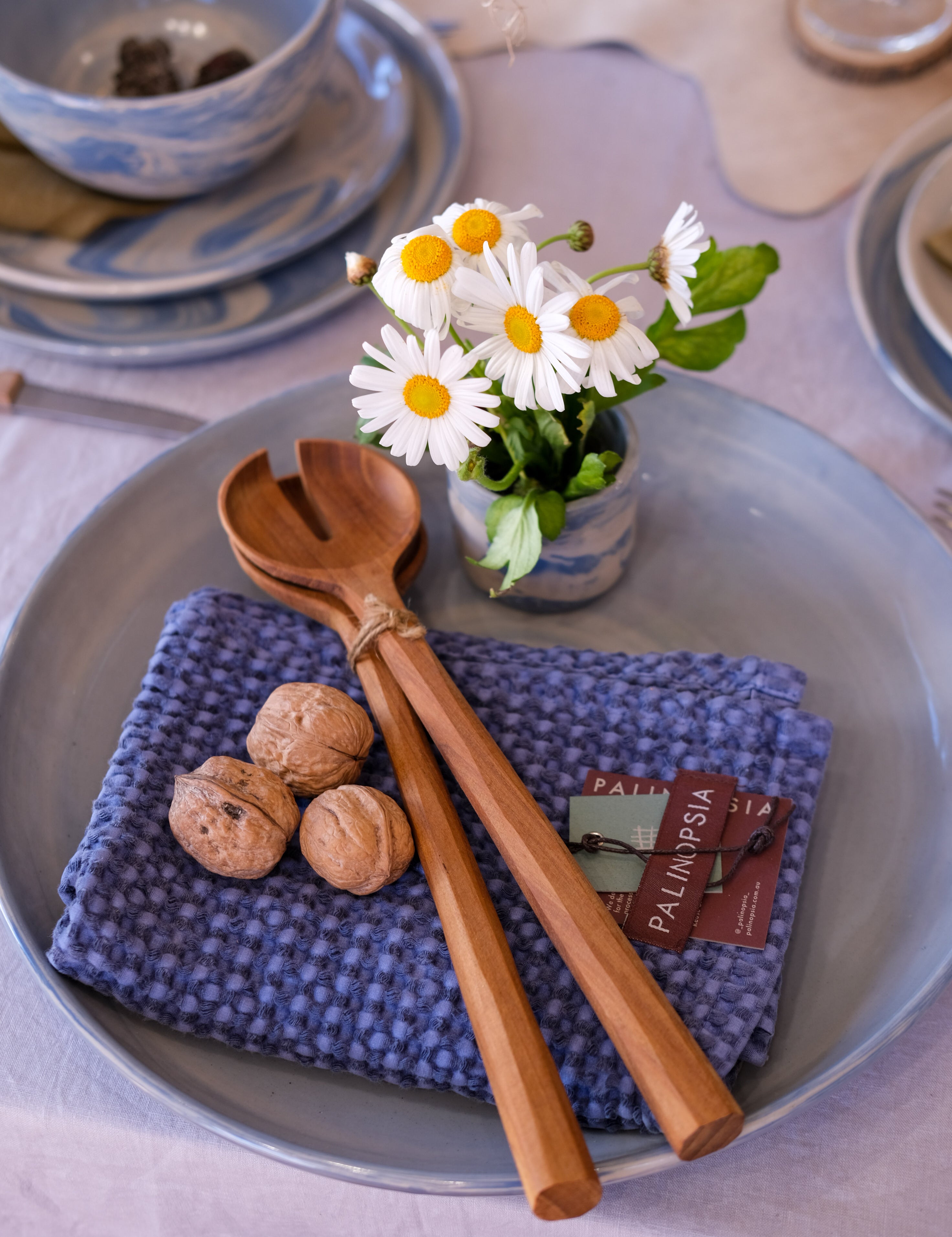 A pair of large teak wooden salad servers on a denim blue waffle tea towel, on a large round ceramic platter. A nicely set dinner table with a light grey tablecloth and blue dinner plates are in the background. 