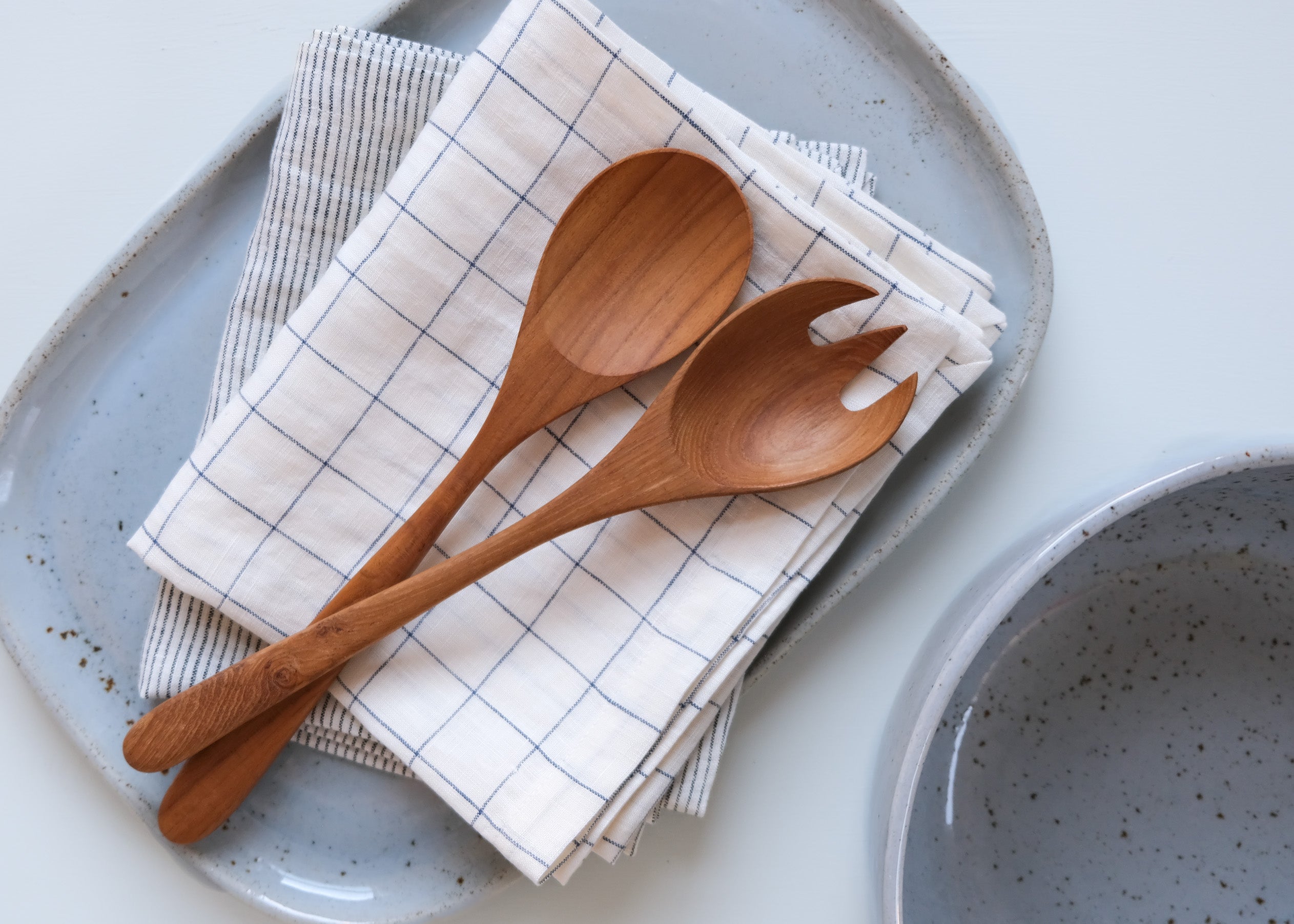 Wooden spoons on a checkered and pinstripe cloth with a Palinopsia ceramic platter and salad bowl in the background.