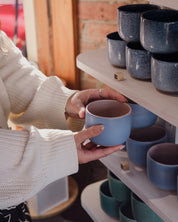 A person in a white knit sweater holds a light blue ceramic mug in a Newcastle ceramic homewares shop, with shelves of similar colourful mugs in the background.