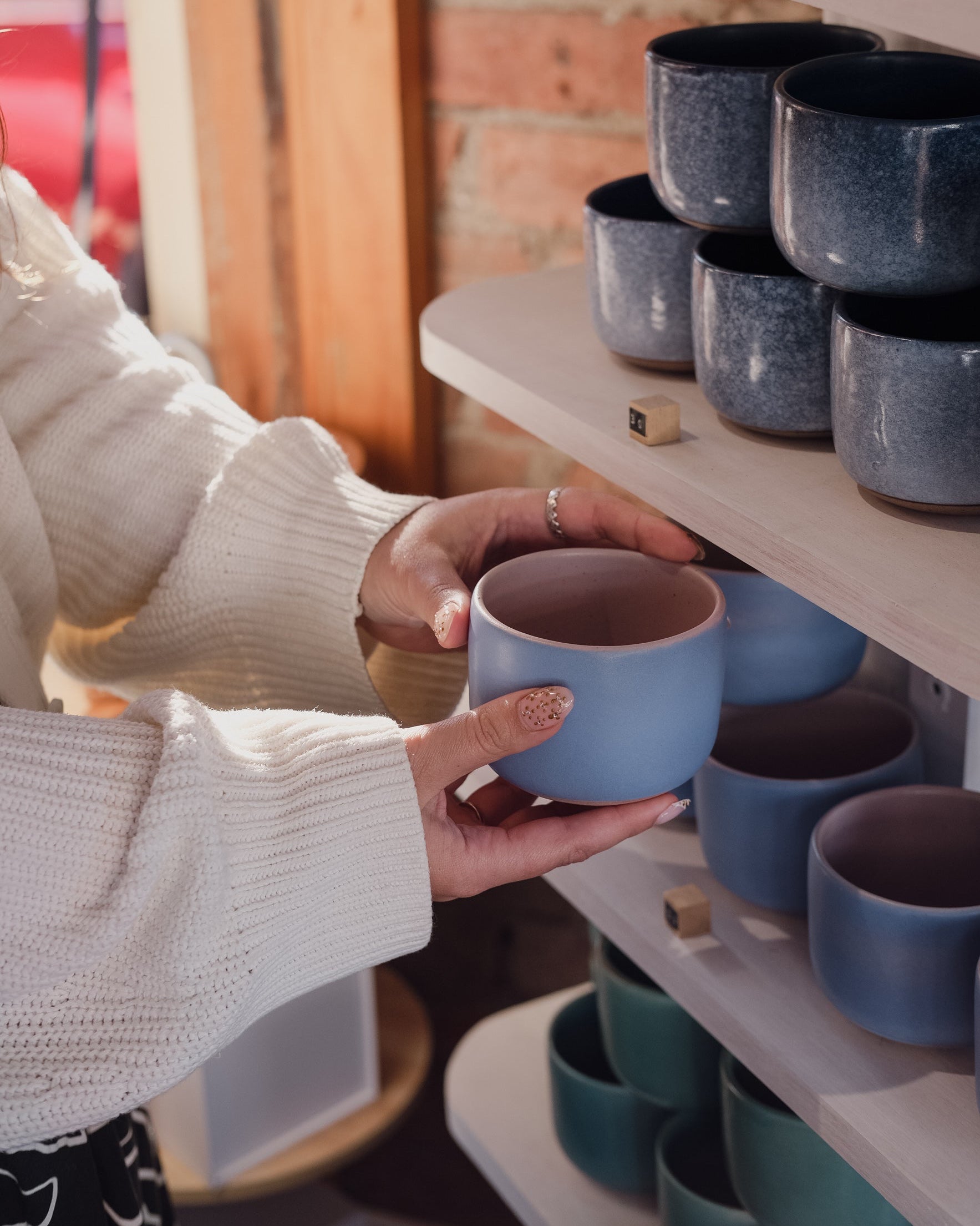 A person in a white knit sweater holds a light blue ceramic mug in a Newcastle ceramic homewares shop, with shelves of similar colourful mugs in the background.