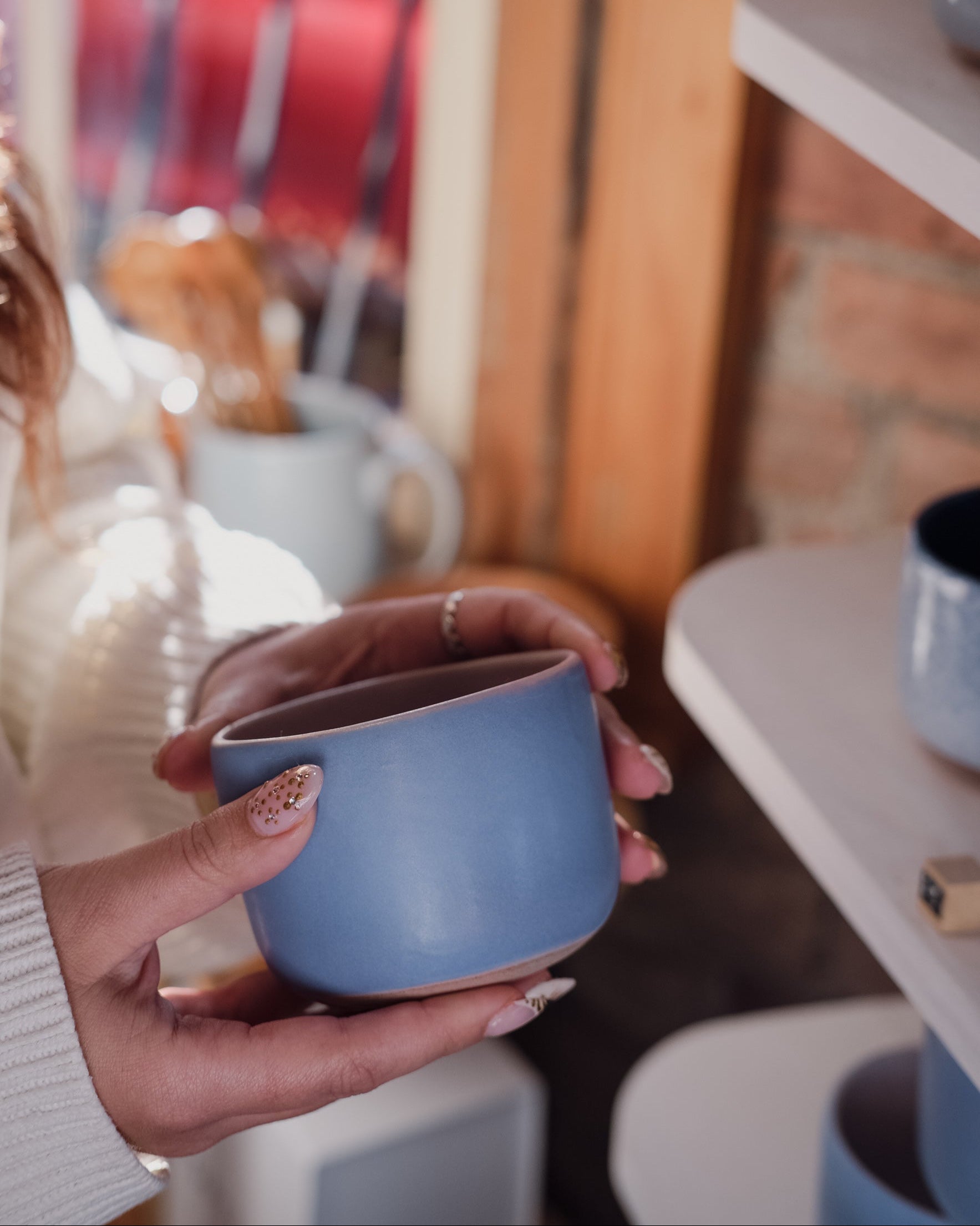 A woman holding a light baby blue ceramic mug with both hands, set against a warm, softly blurred background of shelves and ceramic home decor.