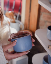 A woman holding a light baby blue ceramic mug with both hands, set against a warm, softly blurred background of shelves and ceramic home decor.
