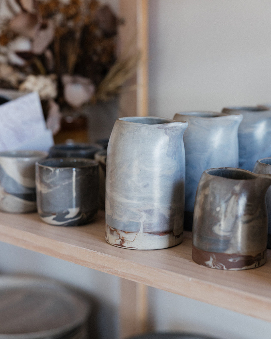 Wooden shelf in a shop with ceramic hand pitchers and coffee cups, out of focus dried flowers decorate the background 