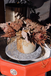 A dried floral arrangement in a textured beige ceramic pot, displayed on a round grey plate resting on a bright orange-red vintage stand up ceramic wheel at Palinopsia Ceramics.