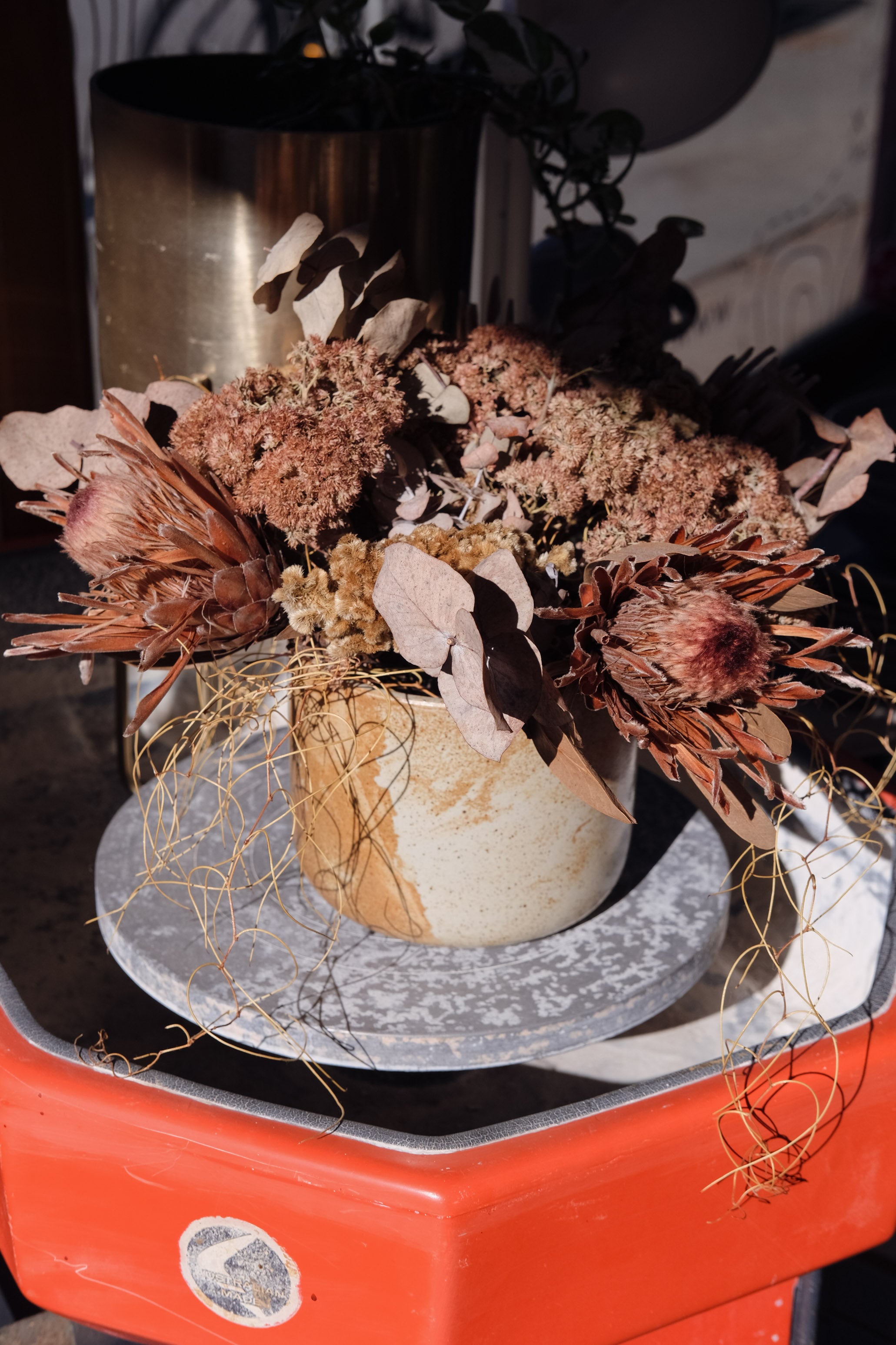 A dried floral arrangement in a textured beige ceramic pot, displayed on a round grey plate resting on a bright orange-red vintage stand up ceramic wheel at Palinopsia Ceramics.