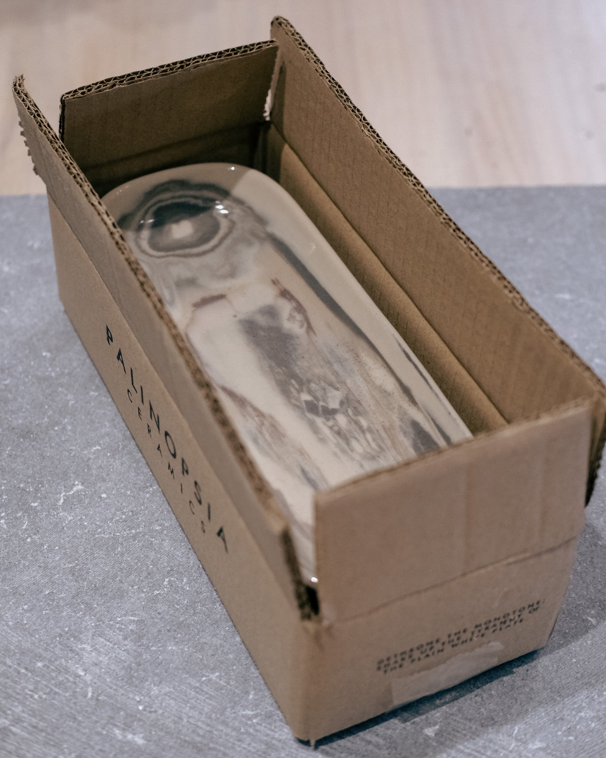 A rectangular glass bottle with a marbled gray and white pattern, partially tucked inside an open brown cardboard box on a gray surface.
