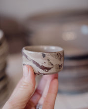 A hand holding a small ceramic ramekin with a brown and beige marble pattern, with a blurred background showing stacks of similar handmade ceramic dishes.