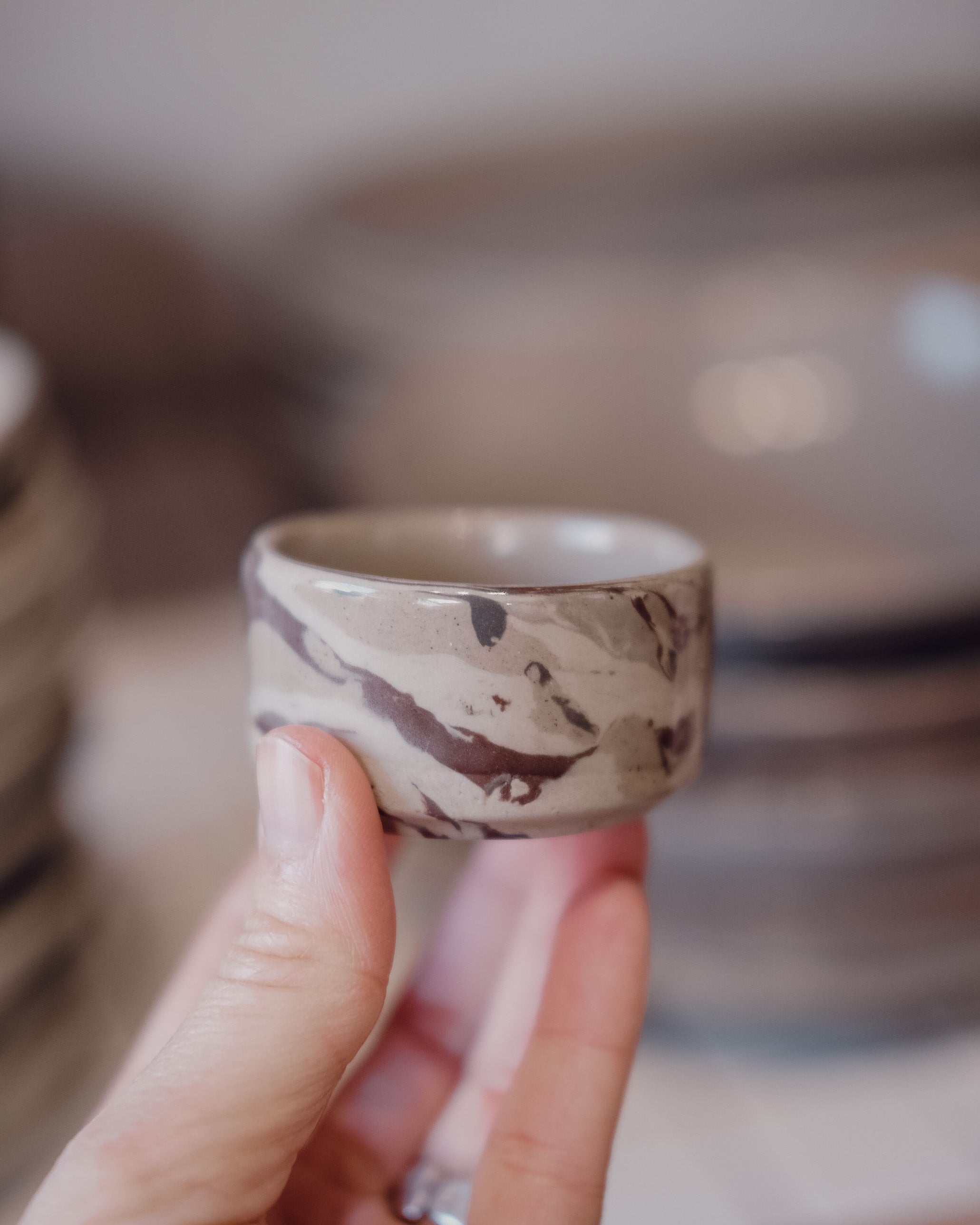 A hand holding a small ceramic ramekin with a brown and beige marble pattern, with a blurred background showing stacks of similar handmade ceramic dishes.