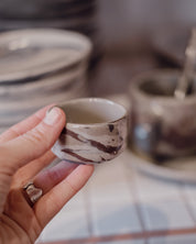 A hand holding a small handmade ceramic cup with a brown and beige marbled pattern, with other ceramic dishes blurred in the background.