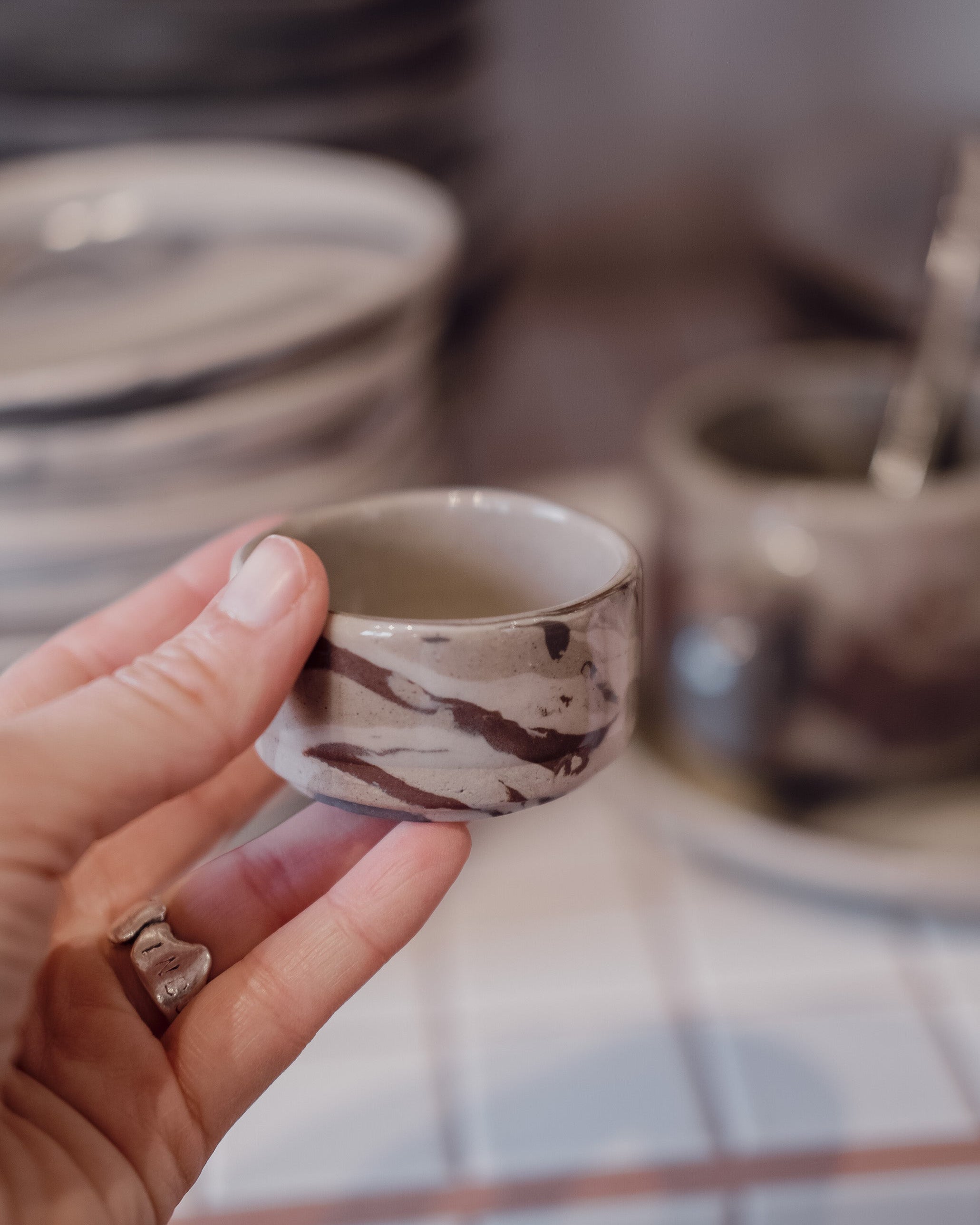 A hand holding a small handmade ceramic cup with a brown and beige marbled pattern, with other ceramic dishes blurred in the background.