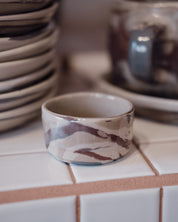 A handmade ceramic small bowl with a marbled pattern of brown, beige, and white, displayed on a white tiled surface with other ceramic dishes blurred in the background.