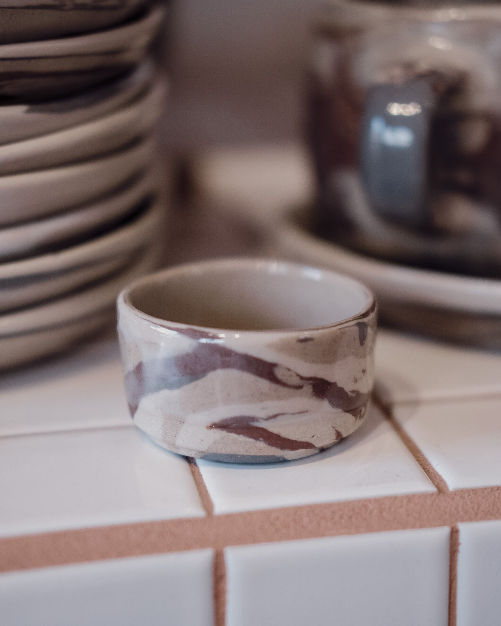 A handmade ceramic small bowl with a marbled pattern of brown, beige, and white, displayed on a white tiled surface with other ceramic dishes blurred in the background.