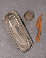 A ceramic rectangle dish tray with a matching small bowl and a wooden cheese knife, arranged on a textured gray surface.