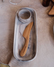 A rectangular ceramic serving dish with a grey, red and white marble pattern, featuring a small matching ramekin inside. A light wood-handled knife with 'PALINOPSIA' engraved on the handle rests on the tray.