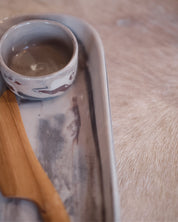 A small handmade ceramic dish with a grey and white marbled pattern sits on a matching rectangular tray, with a wooden knife partially visible, set against a soft beige fur background.