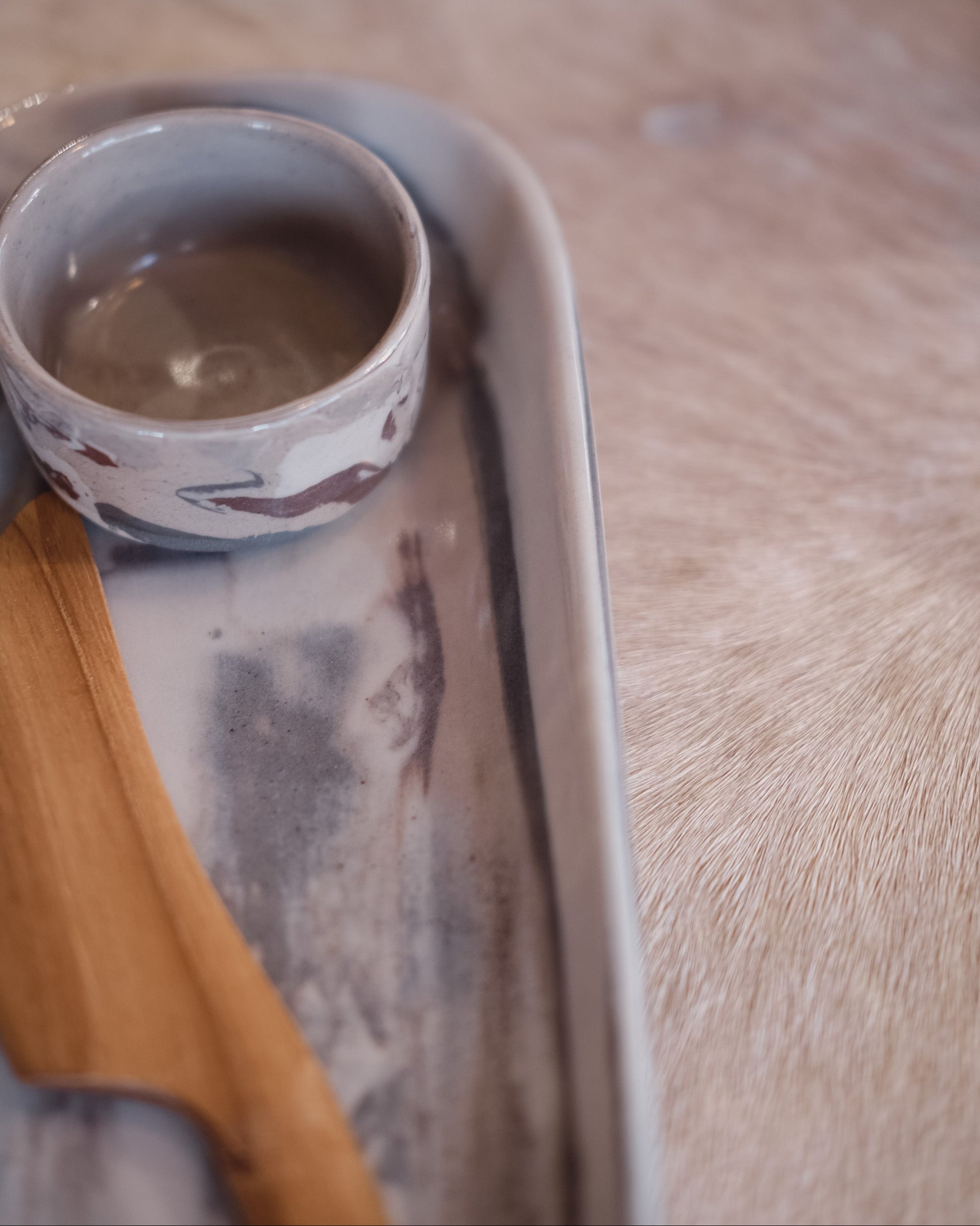 A small handmade ceramic dish with a grey and white marbled pattern sits on a matching rectangular tray, with a wooden knife partially visible, set against a soft beige fur background.