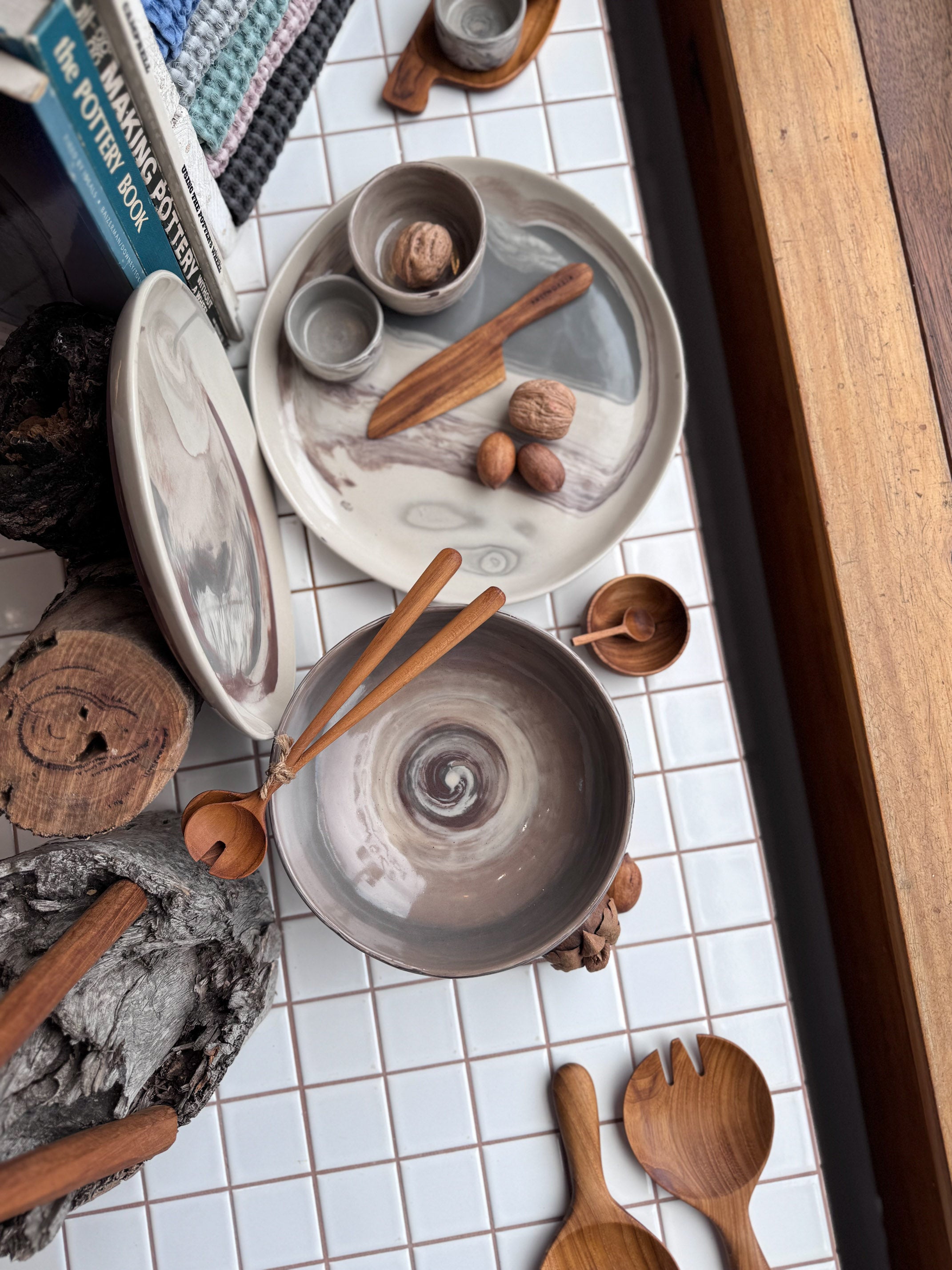 Marbled Ceramic bowls and plates, with teak wooden utensils on a white tiled surface with ceramic books and waffle tea towels in the background.