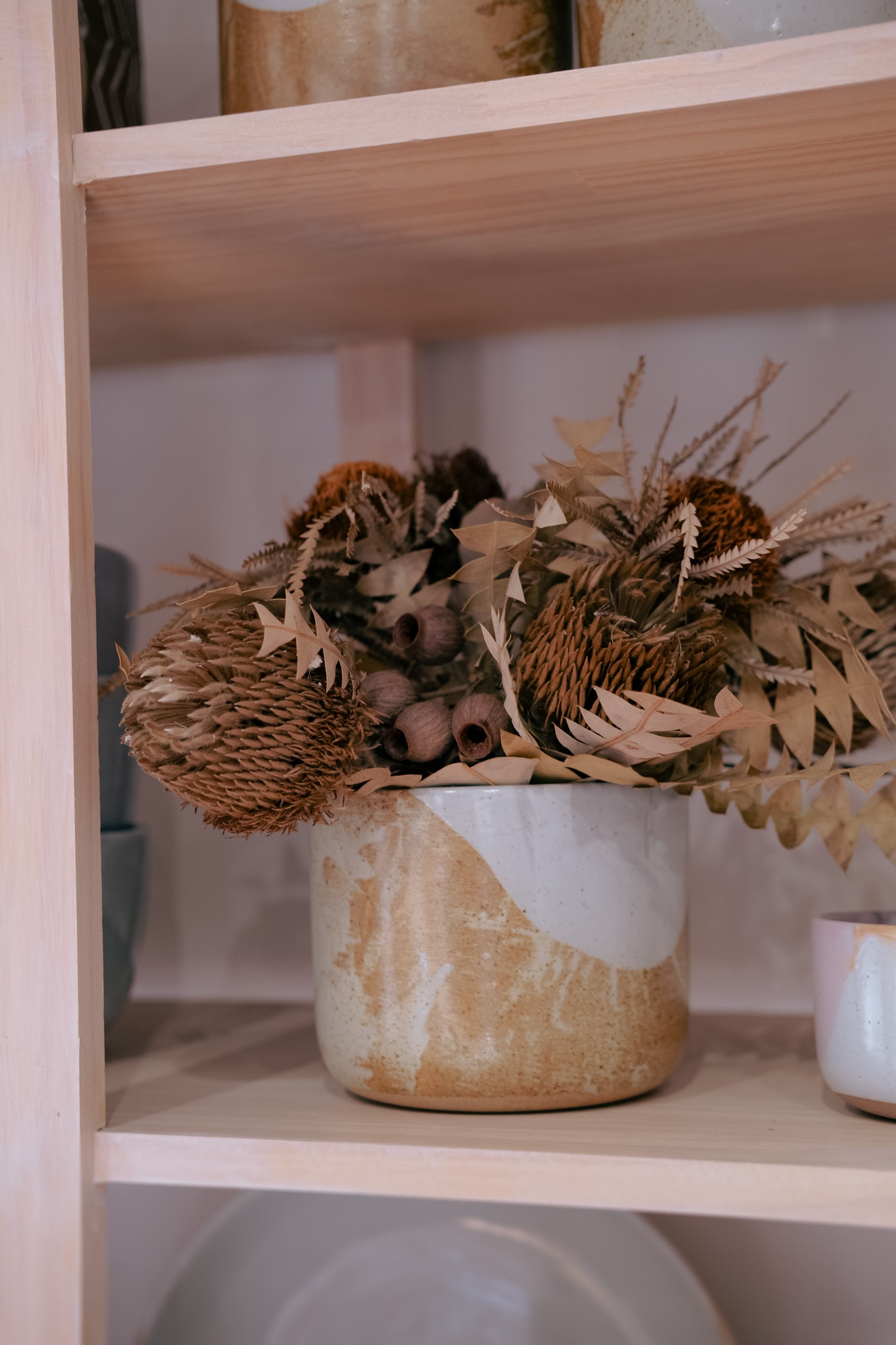 A rustic ceramic pot with a white and beige marbled glaze, filled with dried botanicals including a protea and wheat, displayed on a light wood shelf with colourful pink and chocolate ceramics 